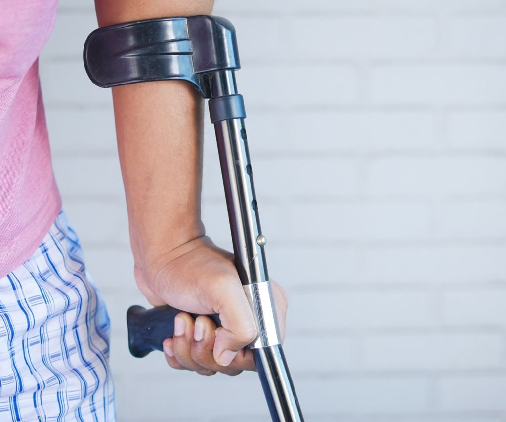 Close-up of a person in a pink shirt using a crutch for support indoors against a brick wall.